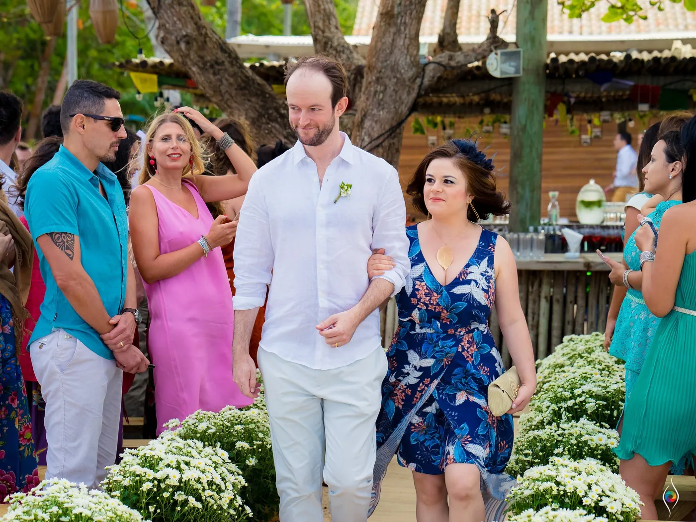 A man in white walks arm in arm with a woman in a blue floral dress up the aisle