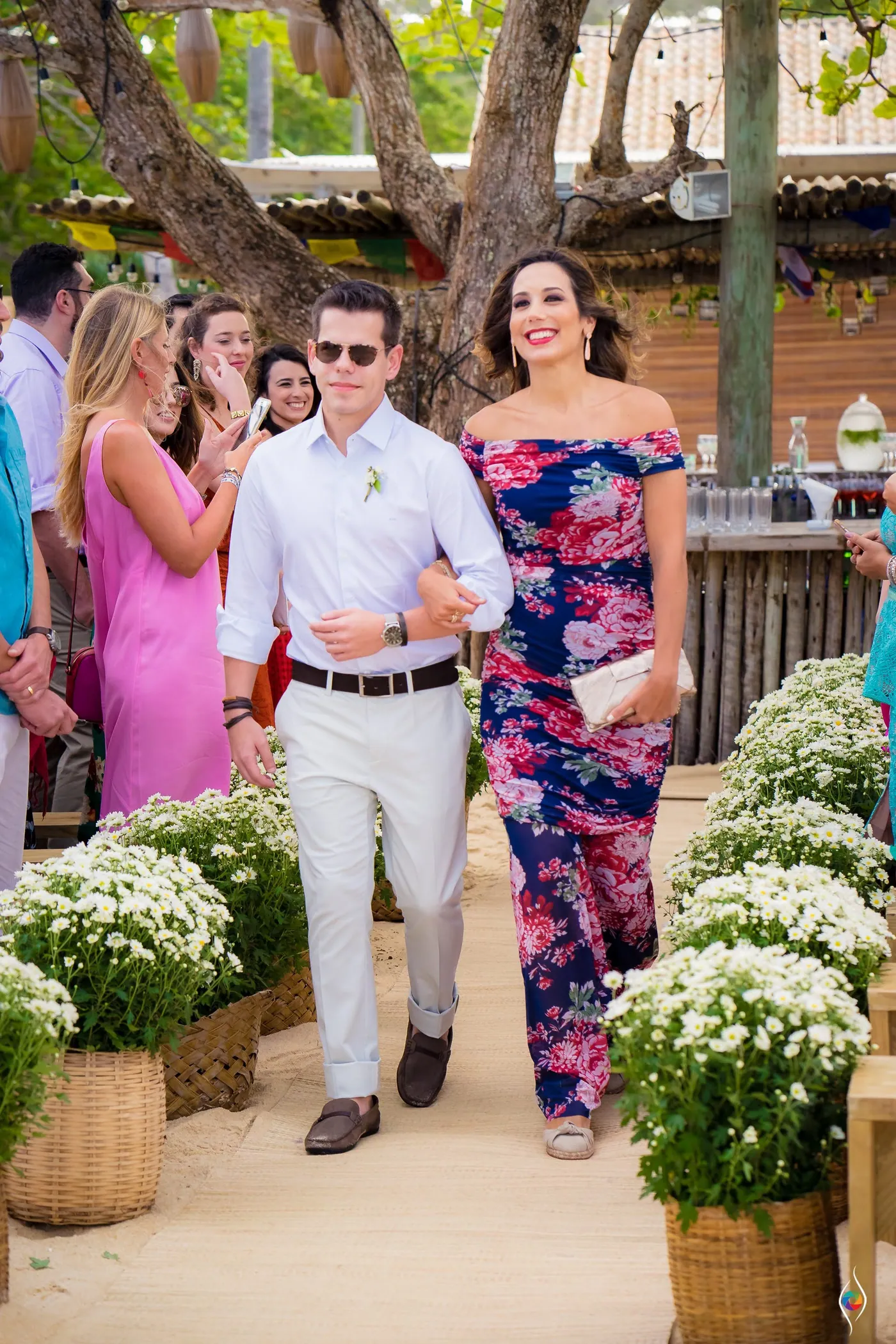 A man in sunglasses and a woman in a navy floral dress walk the sandy aisle past white flower baskets