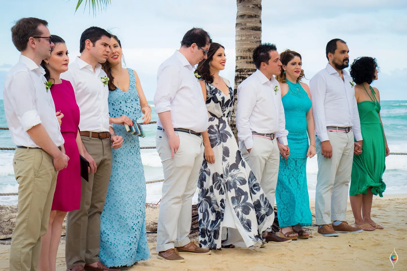 Guests in white, turquoise and green stand in a line on the sand facing the sea
