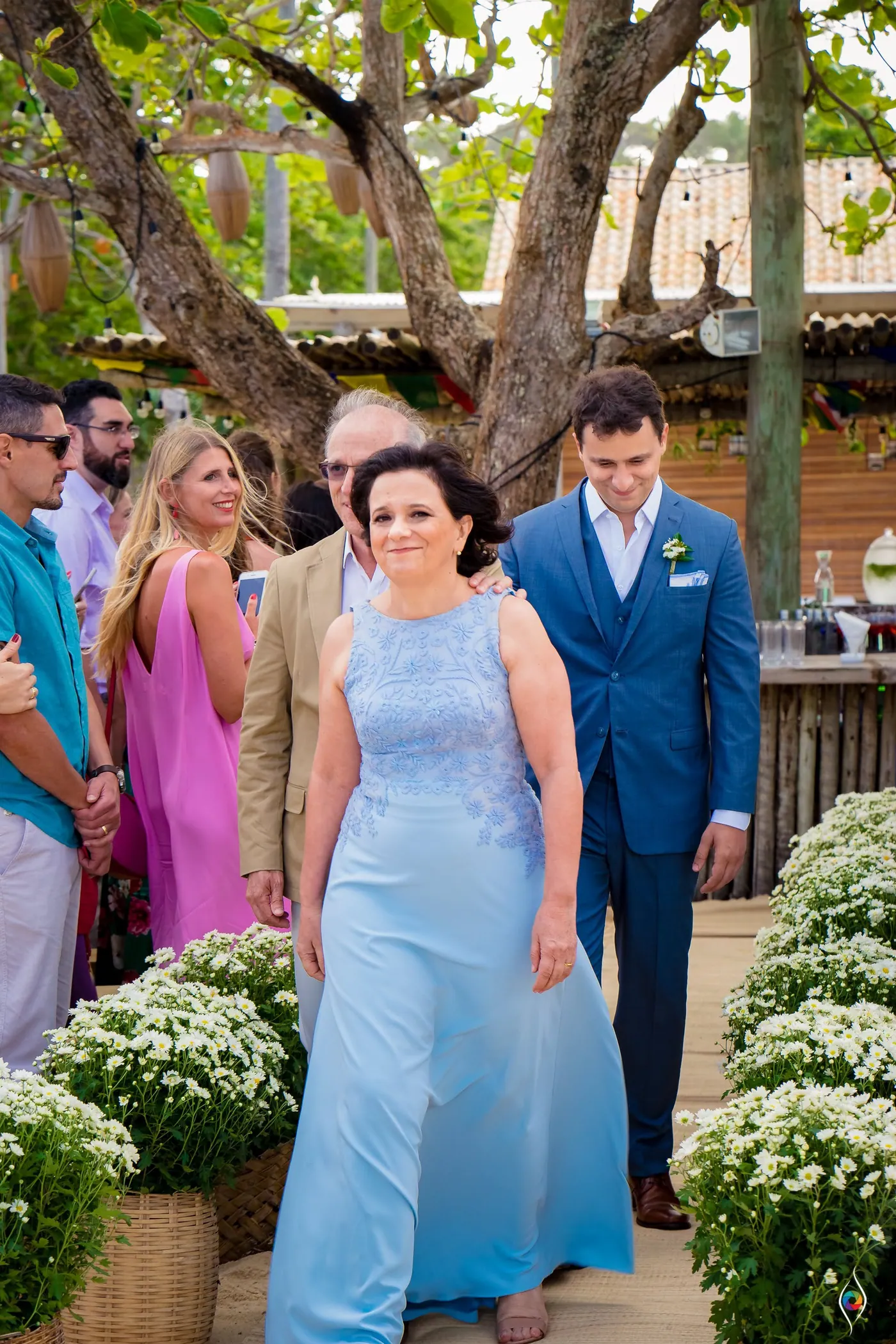 Thiago in his blue suit walks the sandy aisle beside a woman in pale blue, white flowers lining the path