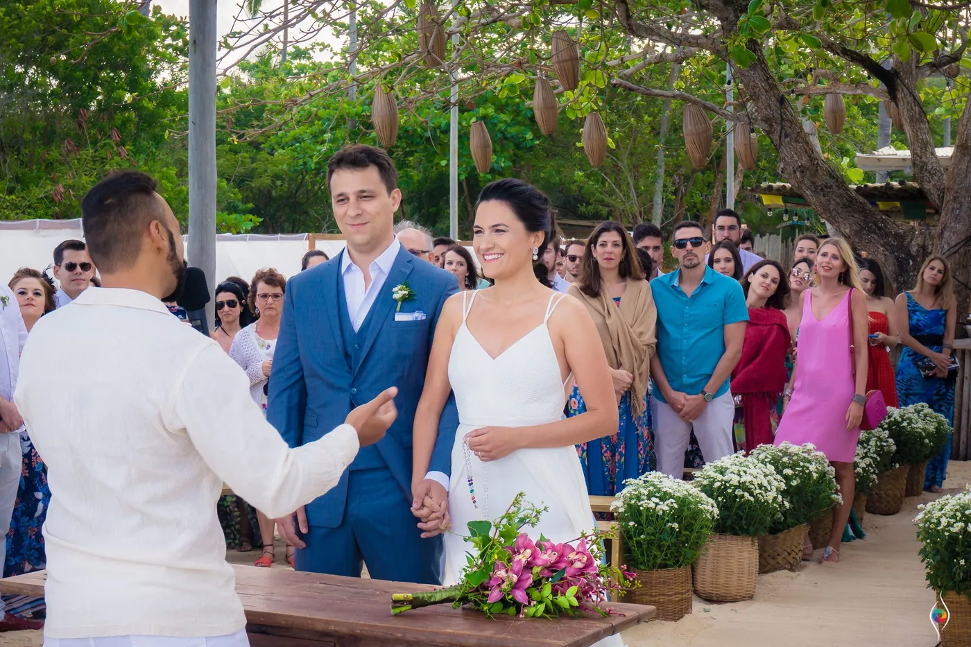 Evy and Thiago hold hands at a rustic wooden table as a man in white linen gestures toward them