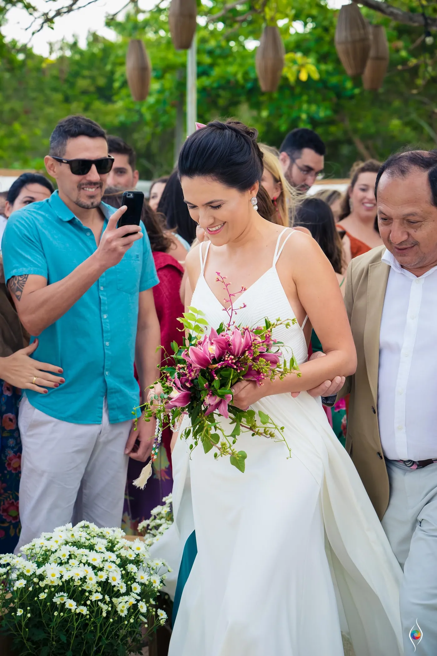 Evy looks down and smiles at her bouquet as she walks, guests holding up phones around her