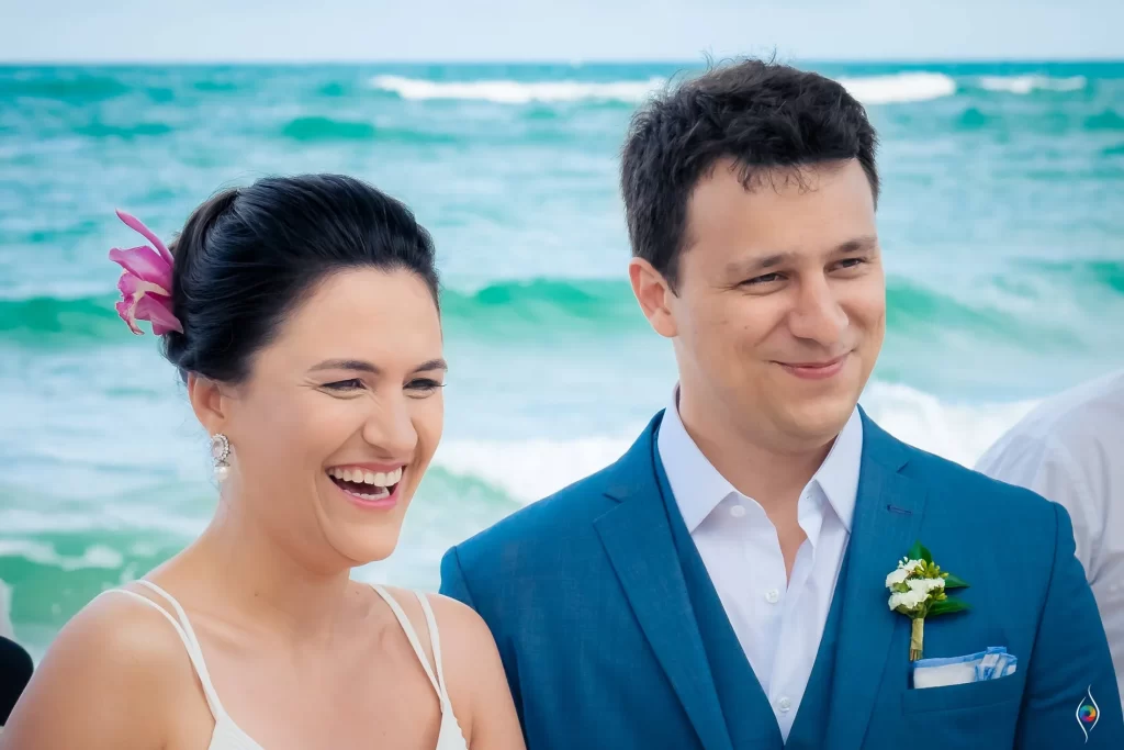Evy and Thiago laugh together with the turquoise sea breaking behind them in Trancoso, Brazil