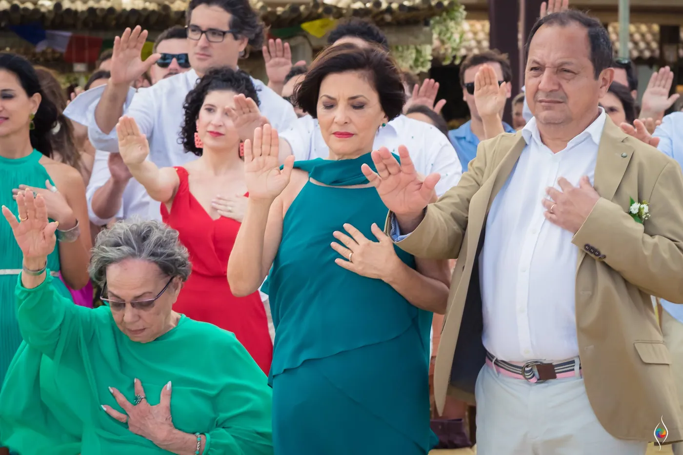 A woman presses a hand to her chest beside a man in a tan jacket as guests raise their palms