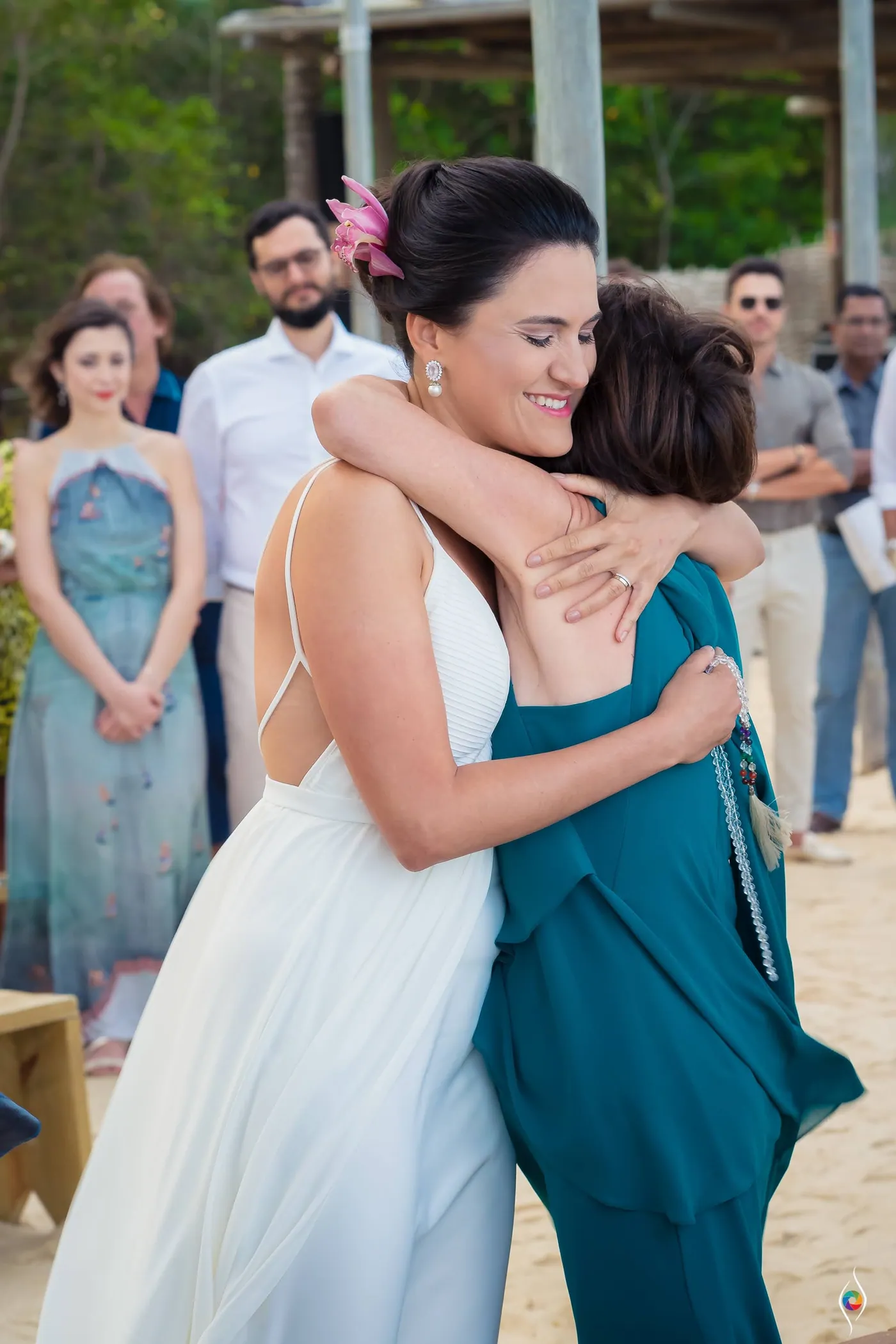 Evy and a woman in teal hold each other tight, her orchid bright against dark hair, guests applauding behind