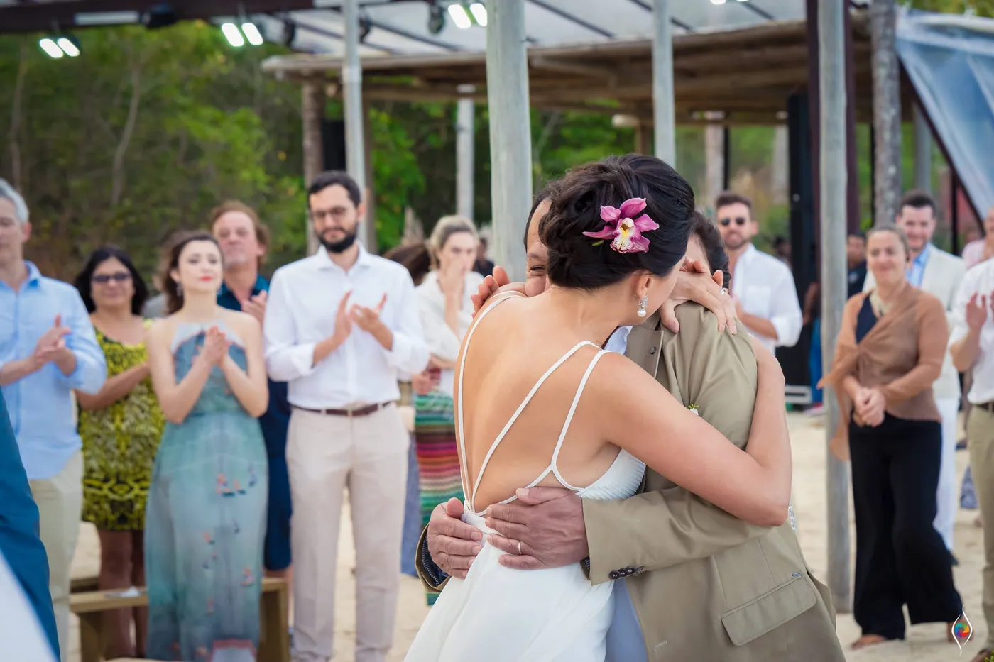 Evy embraces a man in a tan jacket, his hand on her back, guests clapping under the pavilion