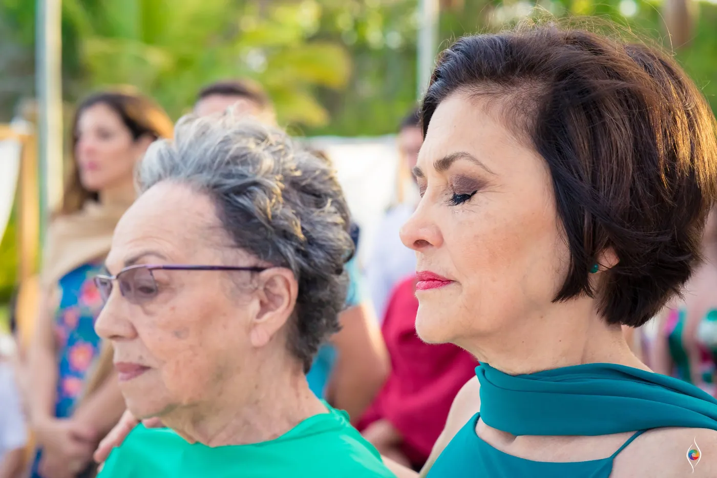 An older woman with her eyes lowered stands beside a woman, both serious and still
