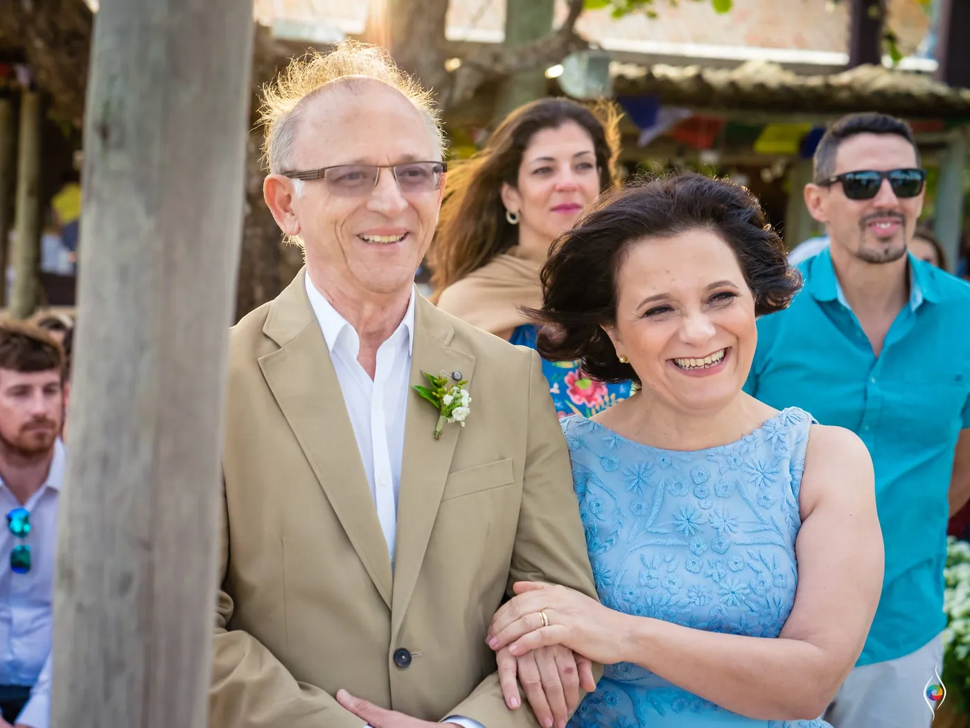 A man in a tan jacket with a boutonniere and a woman in pale blue lace smile arm in arm