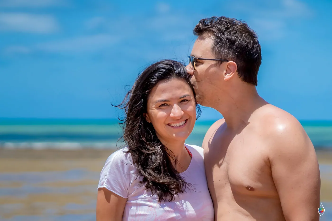 A man kisses a smiling woman on the head with a shallow turquoise sea behind them