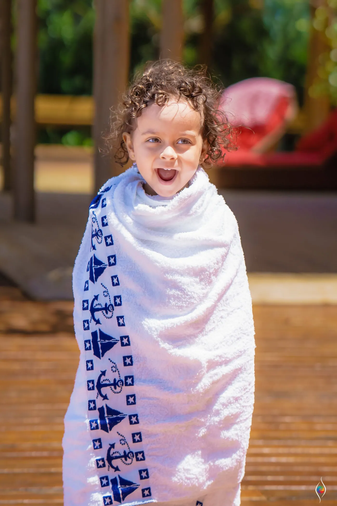 A small child wrapped in a blue anchor-print towel grins on a wooden deck