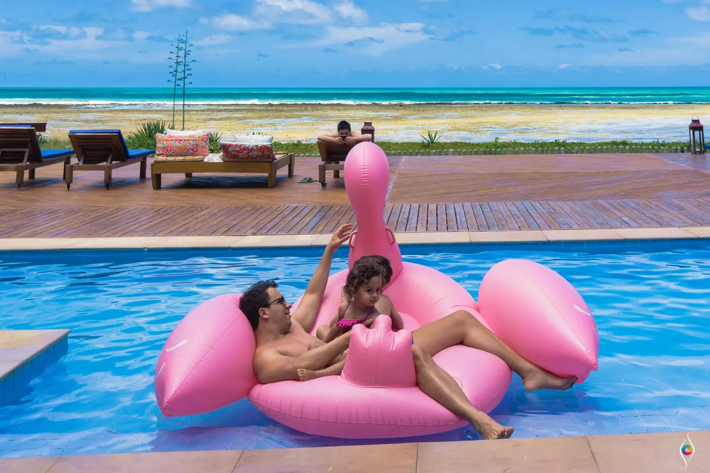 Two people lounge on a giant pink flamingo float in a pool with the sea beyond the deck
