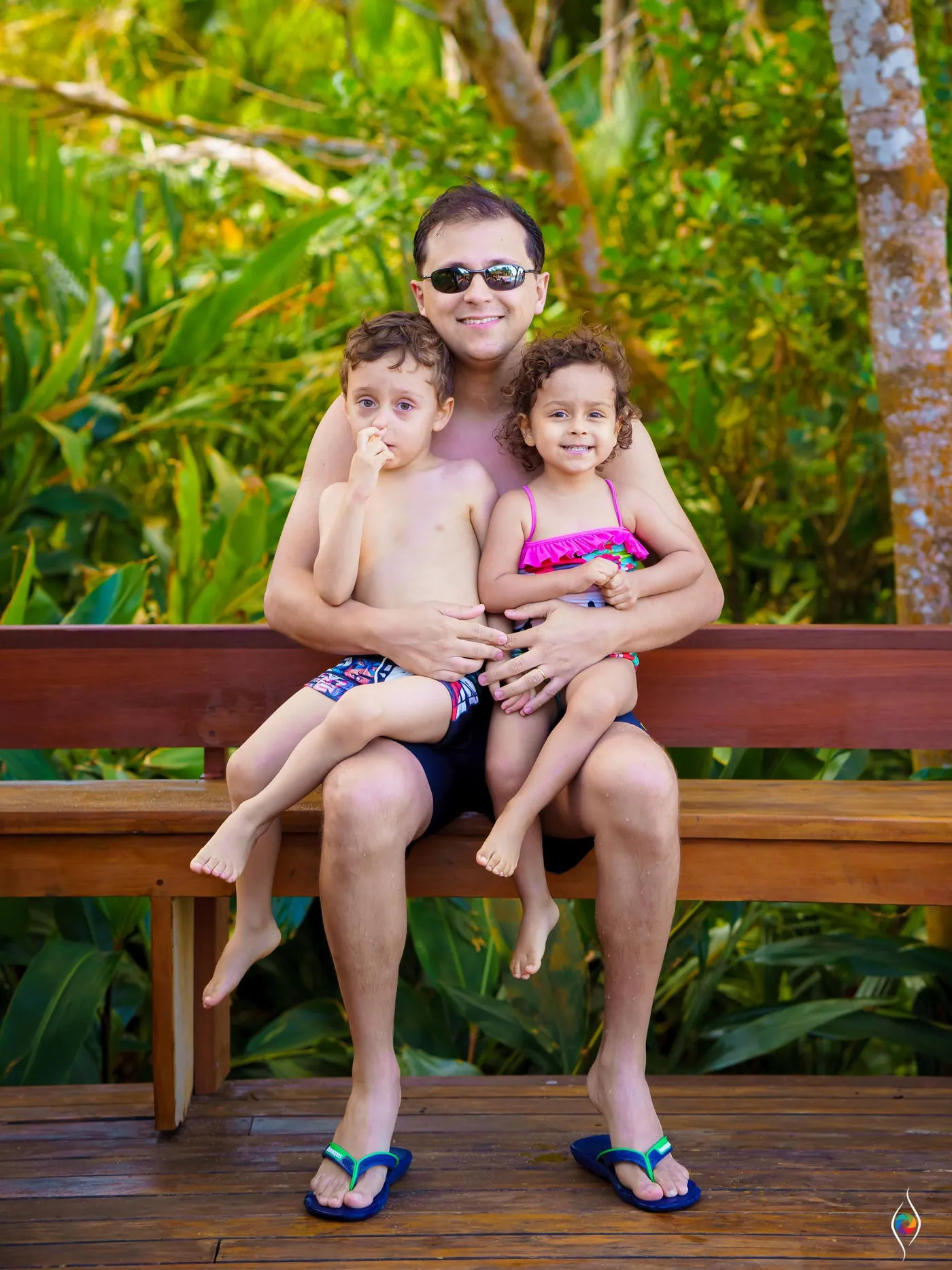 A man in sunglasses sits on a bench with two small children on his lap in a leafy garden