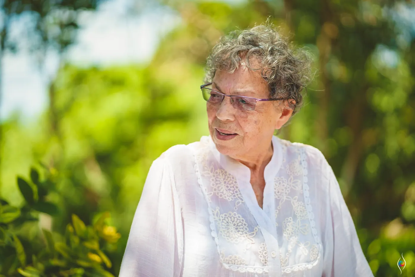 An older woman in a white embroidered blouse looks off to the side among green garden leaves