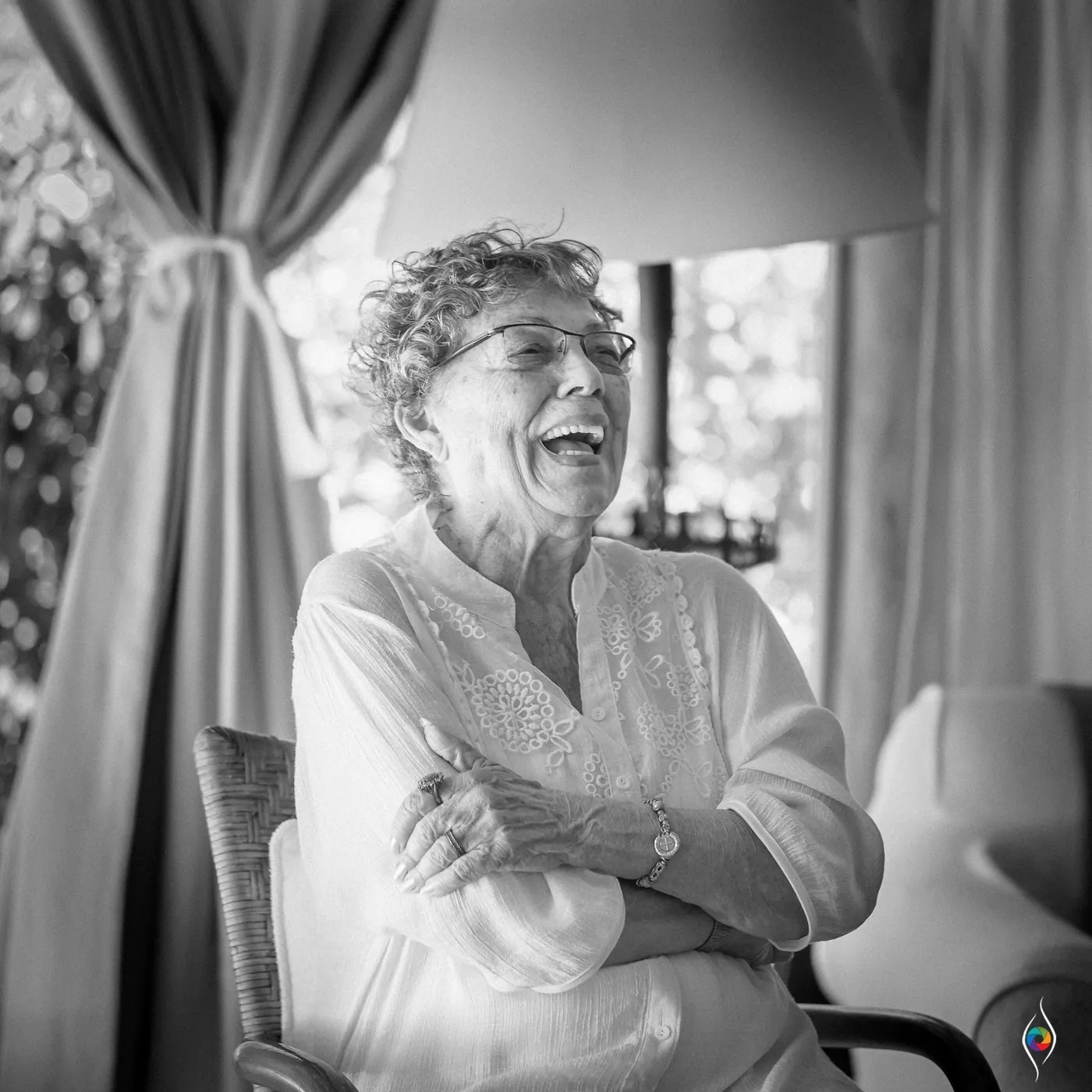 An older woman laughing with her arms folded, seated beside a curtain in black and white