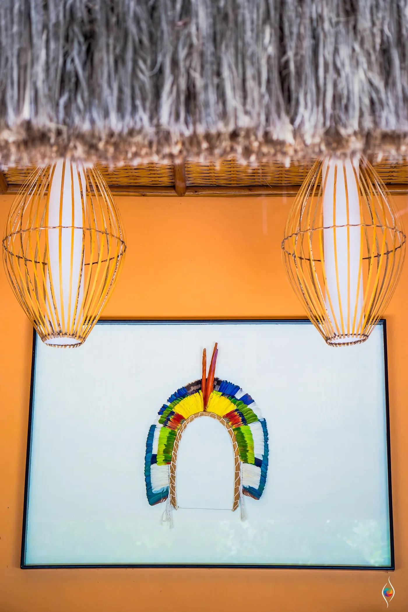 Feathered headdress artwork in a white frame beneath two woven lamps and a thatch roof