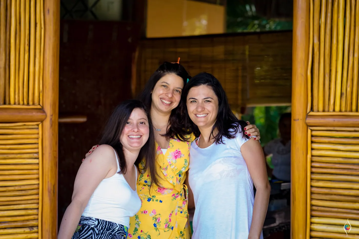 Three women pose arm in arm in a bamboo doorway, the one in the middle wearing yellow florals