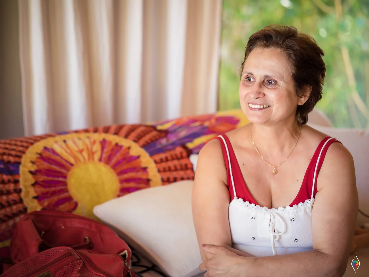 An older woman with short hair smiles from a sofa heaped with a bright sun-print cushion