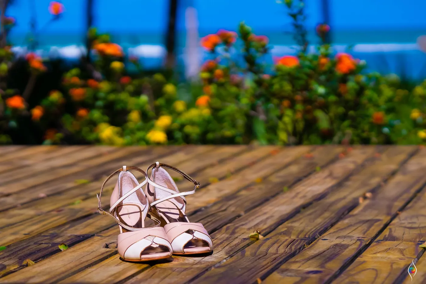 Pale pink sandals sit on a wooden deck with orange flowers and the sea blurred behind