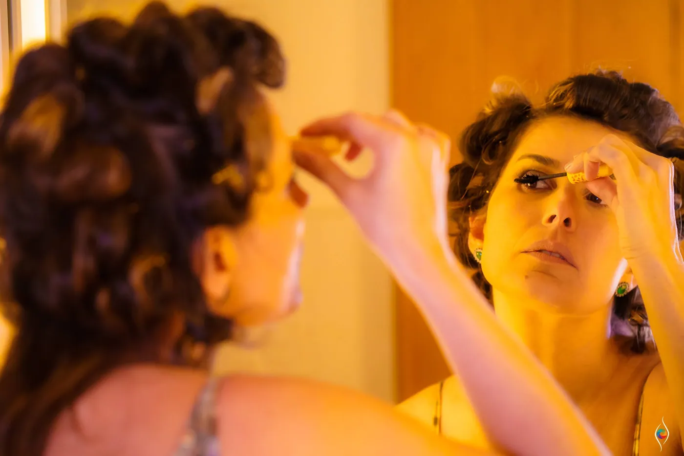 A makeup artist applies eye makeup to a woman with a curled updo in warm window light