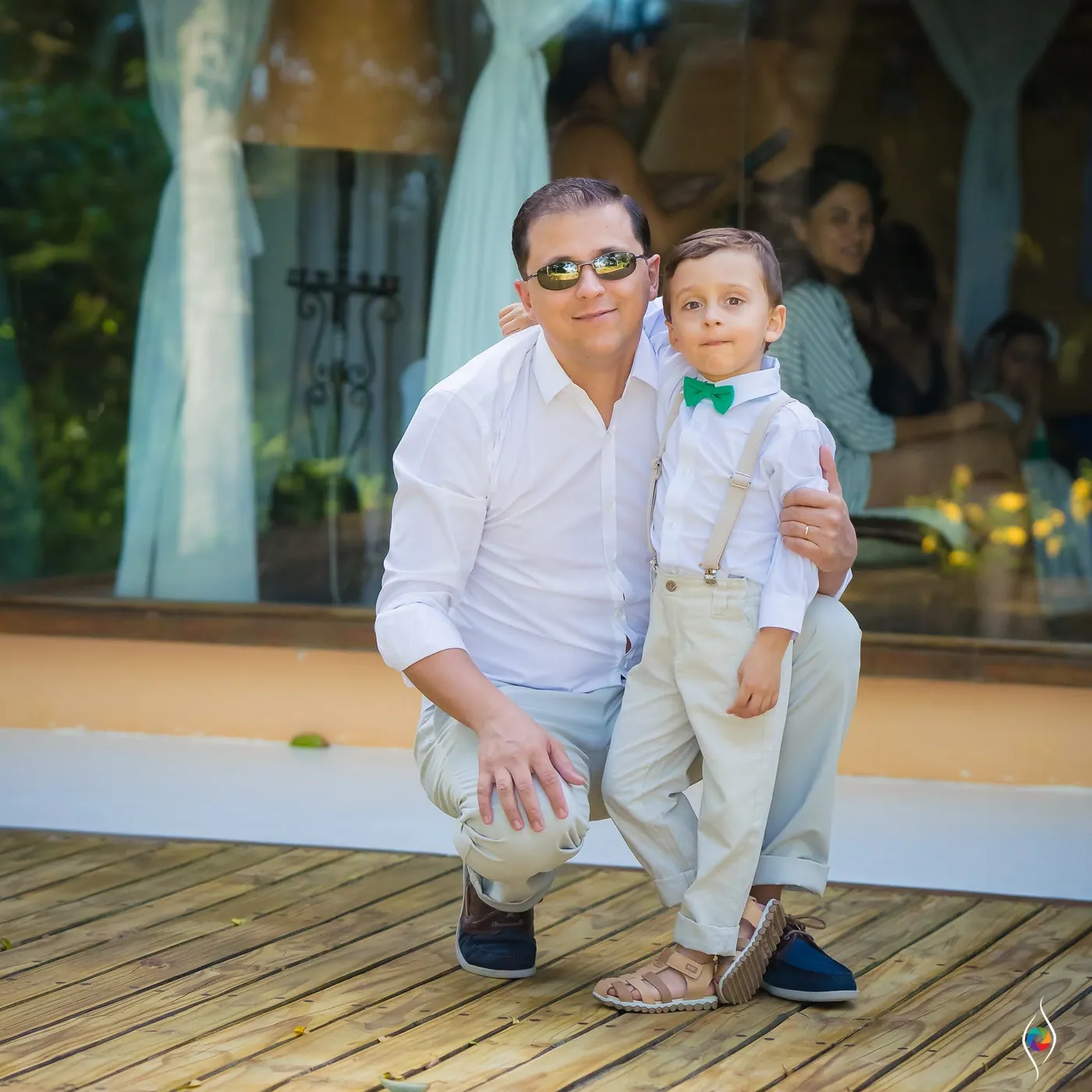 A man in sunglasses crouches on a deck beside a small boy in suspenders and a green bow tie