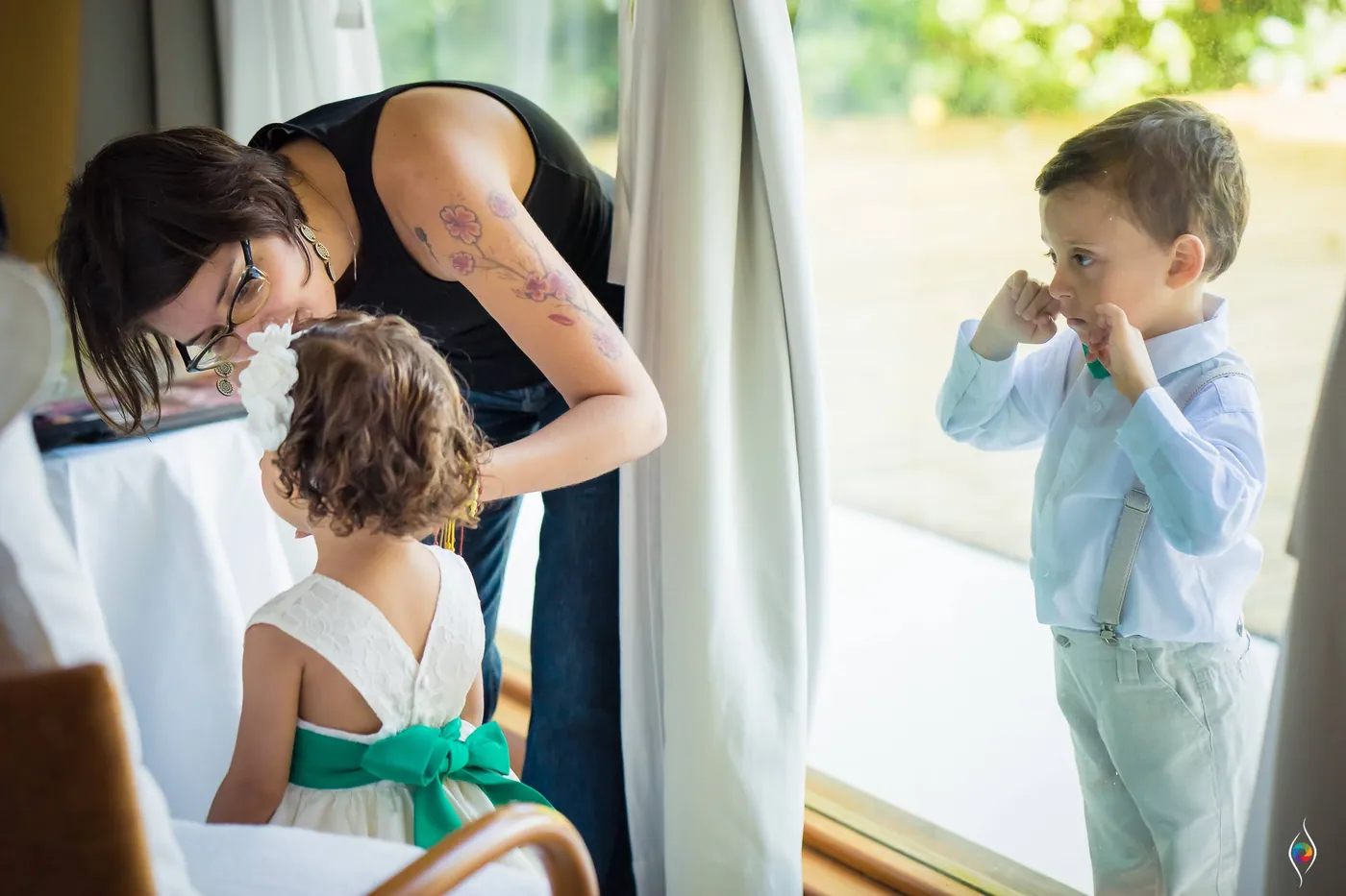 A woman fastens the green sash of a flower girl's dress as a small boy rubs his eye