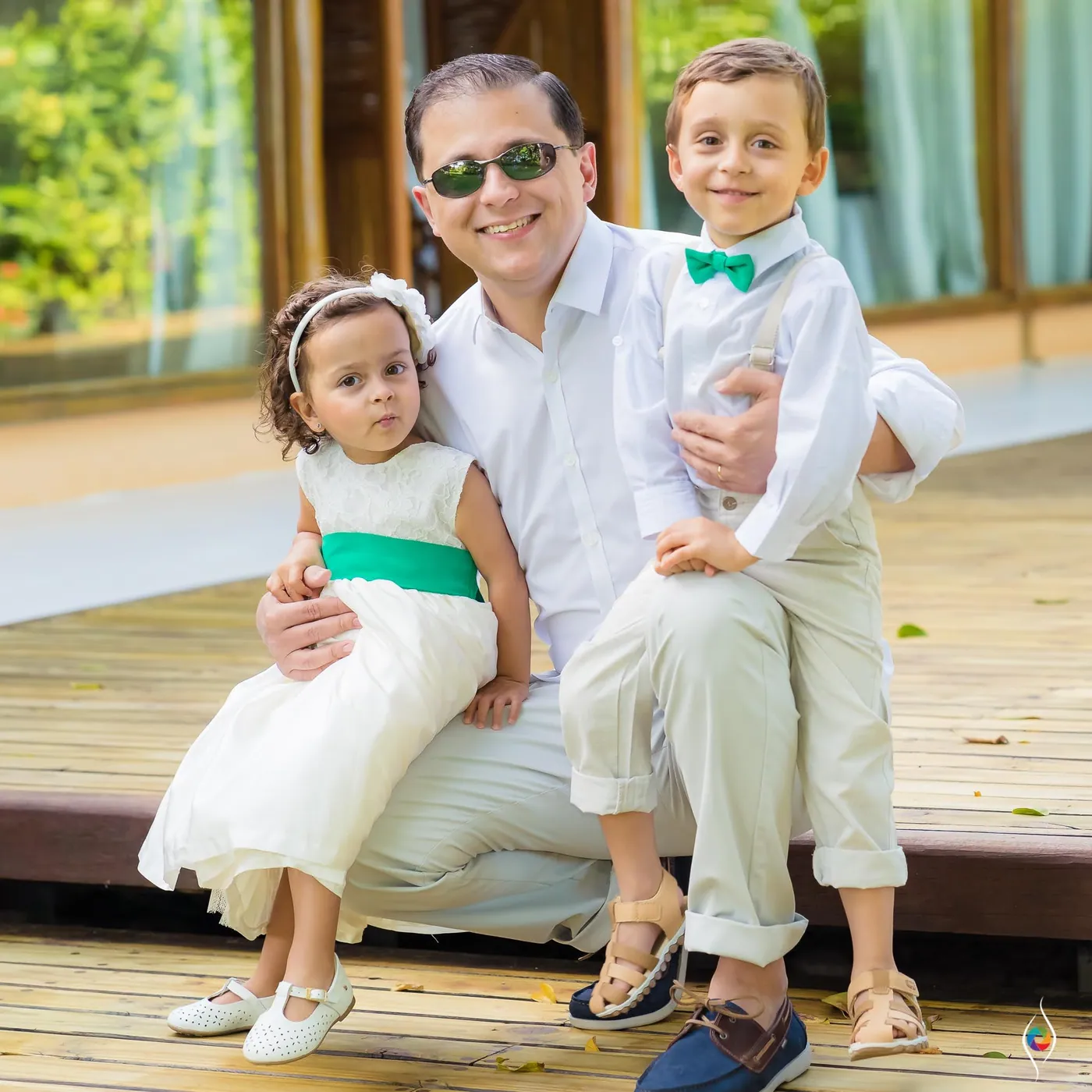 A man sits with a flower girl in white and a boy in a green bow tie on his knees
