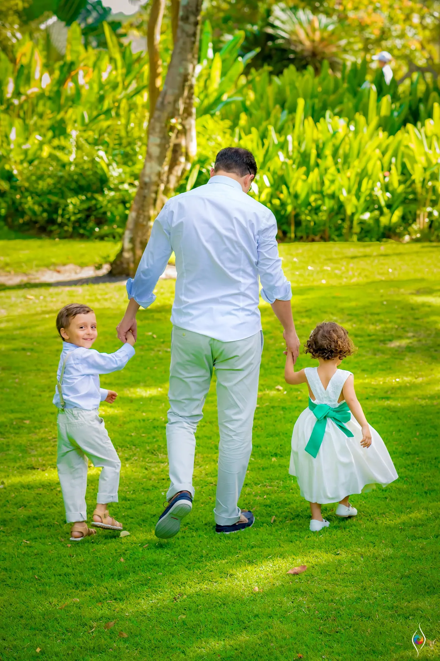 A man walks across a sunny lawn holding hands with a small boy and a girl in a green sash