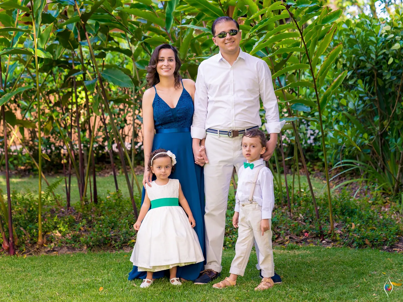 A family of four stands on a lawn among tropical plants, the two children dressed for the wedding