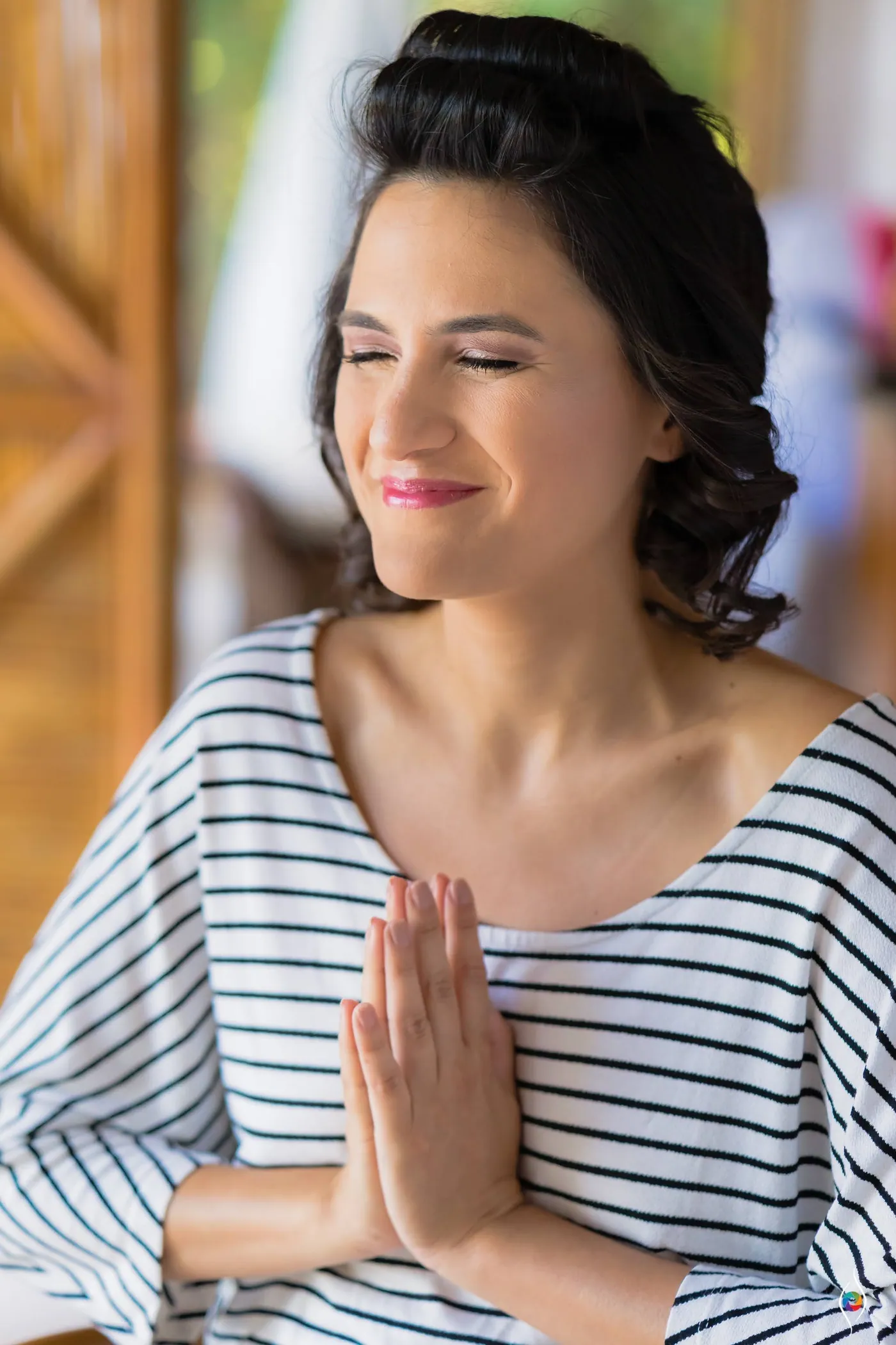 Evy presses her palms together and shuts one eye with a grin, wearing a striped top