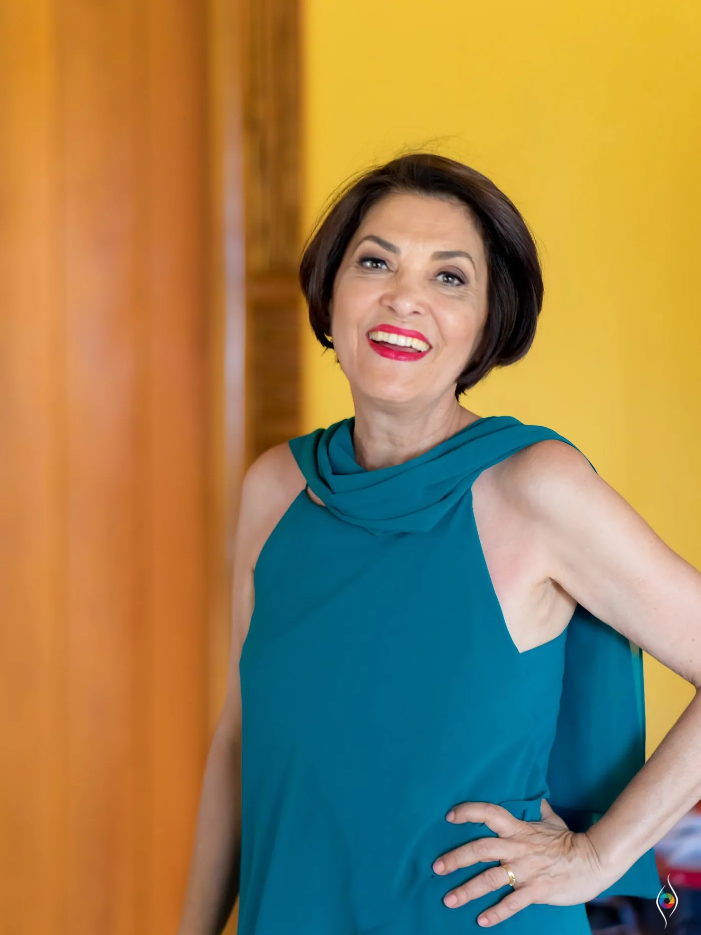 A woman in a teal top poses against a yellow wall with a hand on her hip