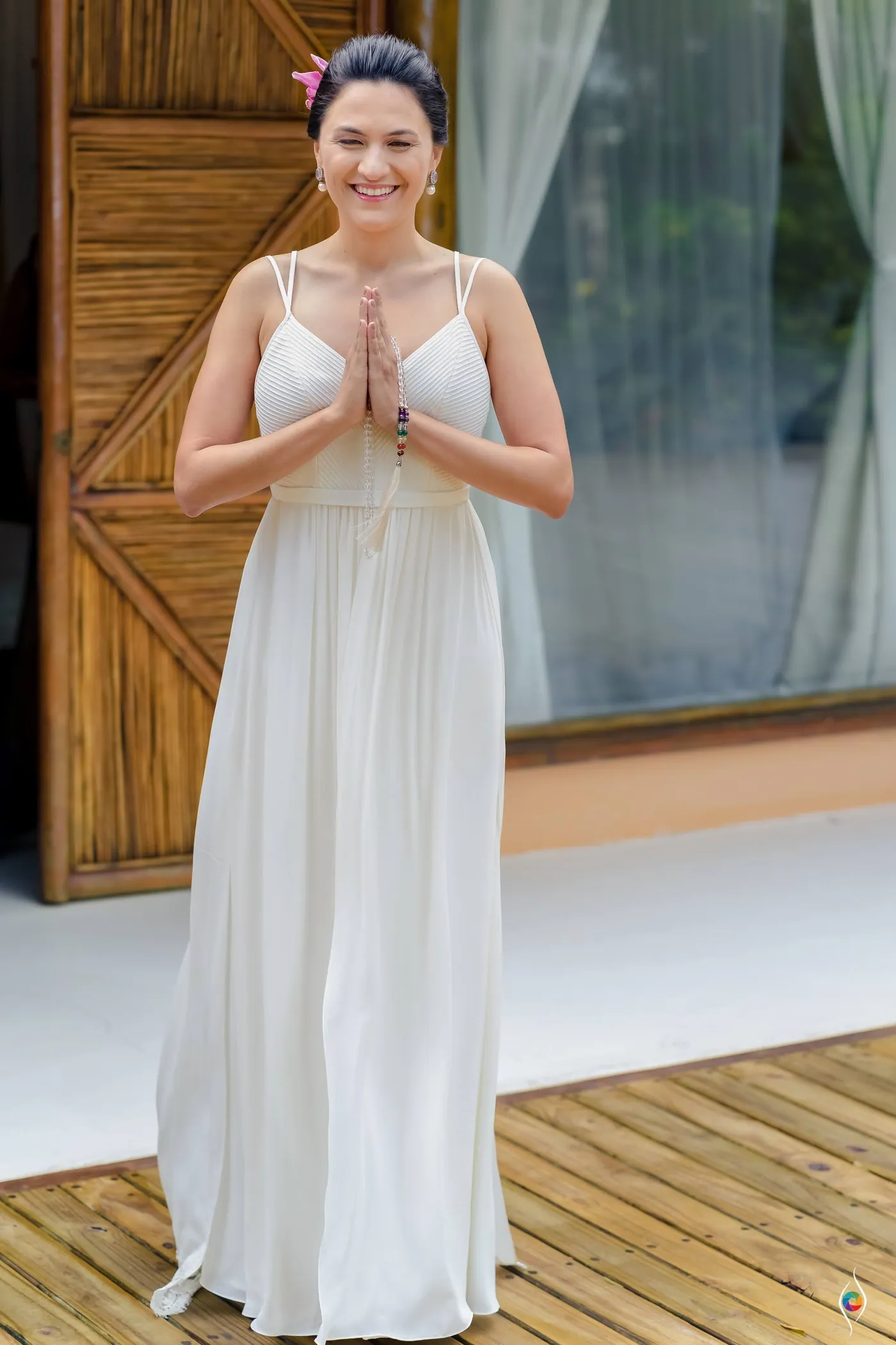 Evy stands on a wooden deck in her long white dress with her palms pressed together