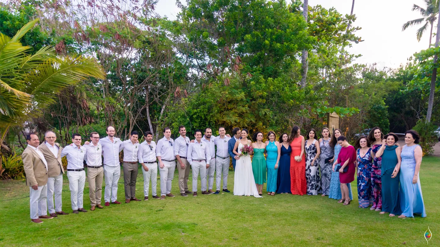 Wide group portrait of the wedding party lined up on a green lawn beneath tall trees