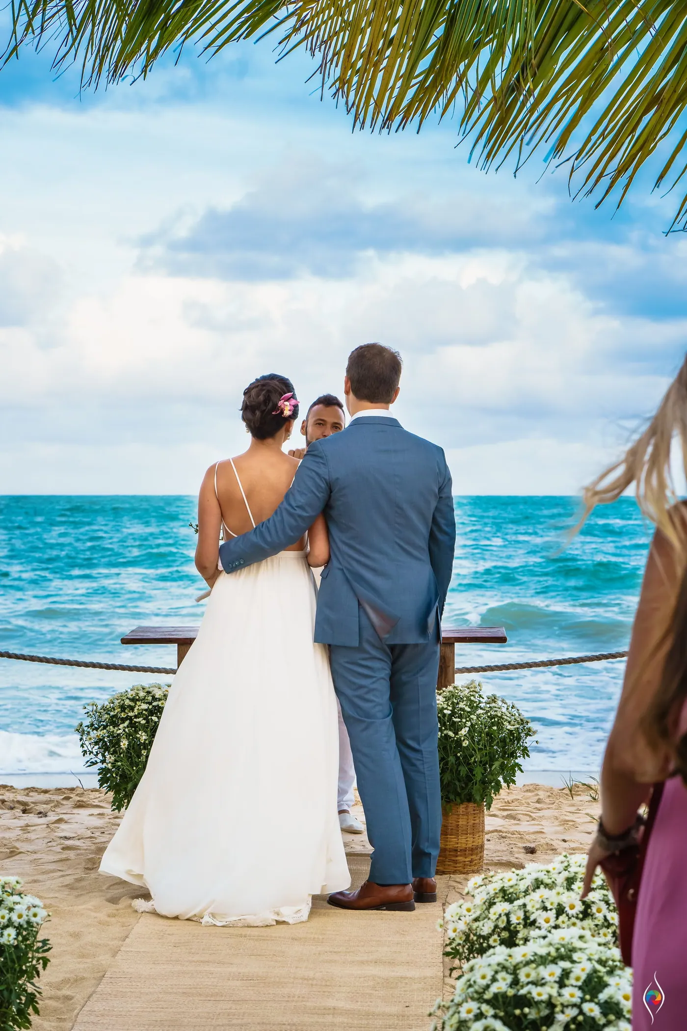 Evy and Thiago stand with their backs to the camera facing the sea, tall white flowers beside them