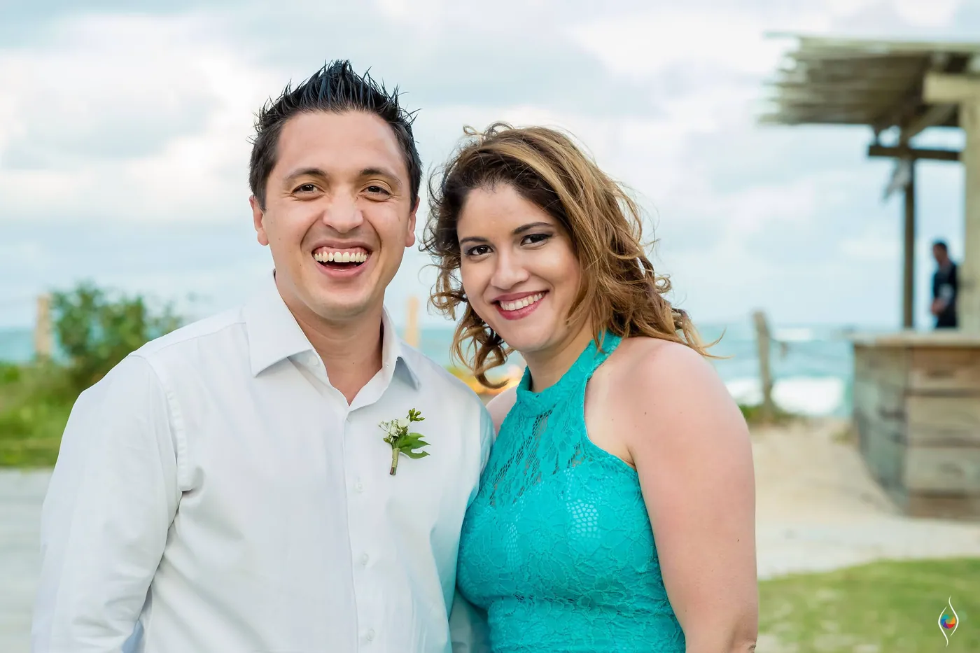 A man laughs with a woman in a teal lace dress, the sea and a thatched bar behind them