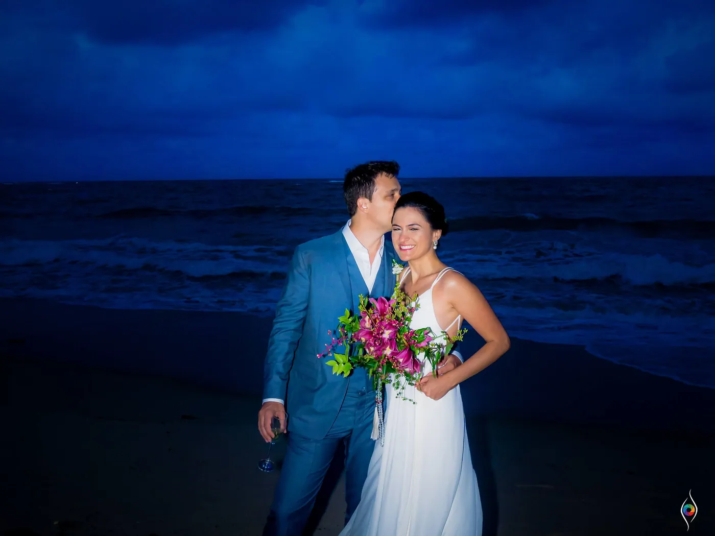 Evy and Thiago stand on the beach at dusk, he kisses her cheek, her bright bouquet in hand