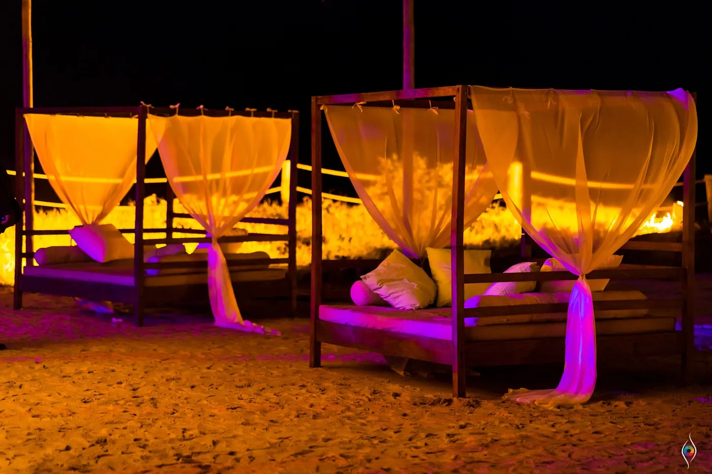 Beach daybeds draped with sheer canopies glow orange and pink on the sand at night