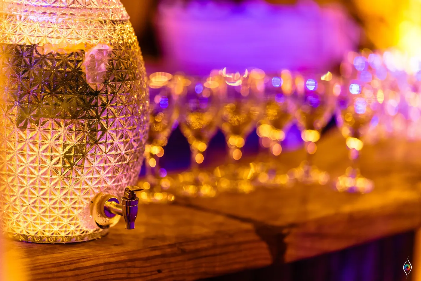 A patterned glass dispenser and rows of champagne flutes glow purple on a bar table
