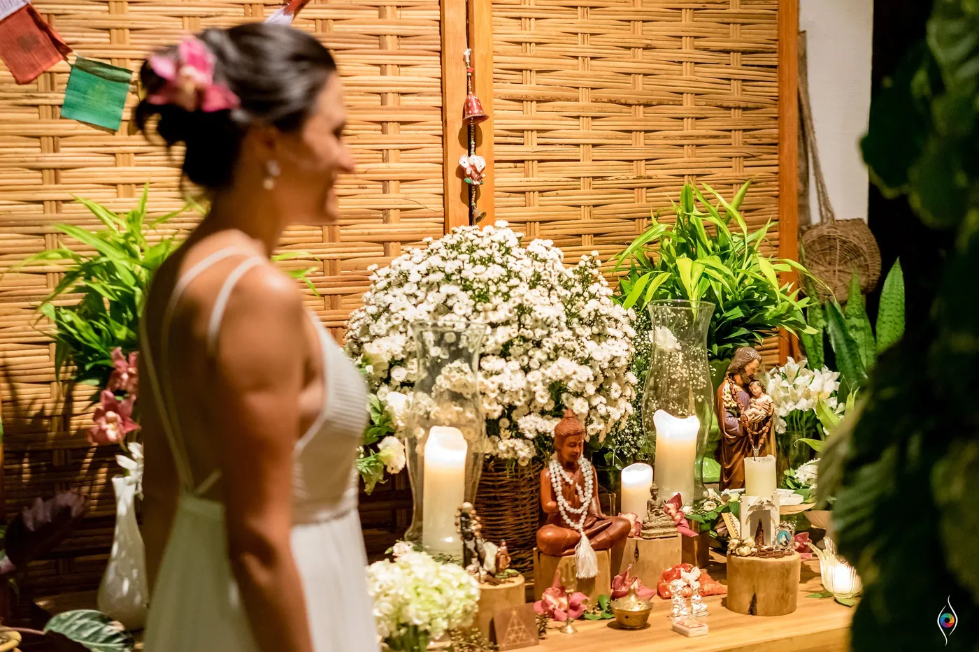Evy stands in profile beside a table of candles, white daisies and small figurines before a woven wall