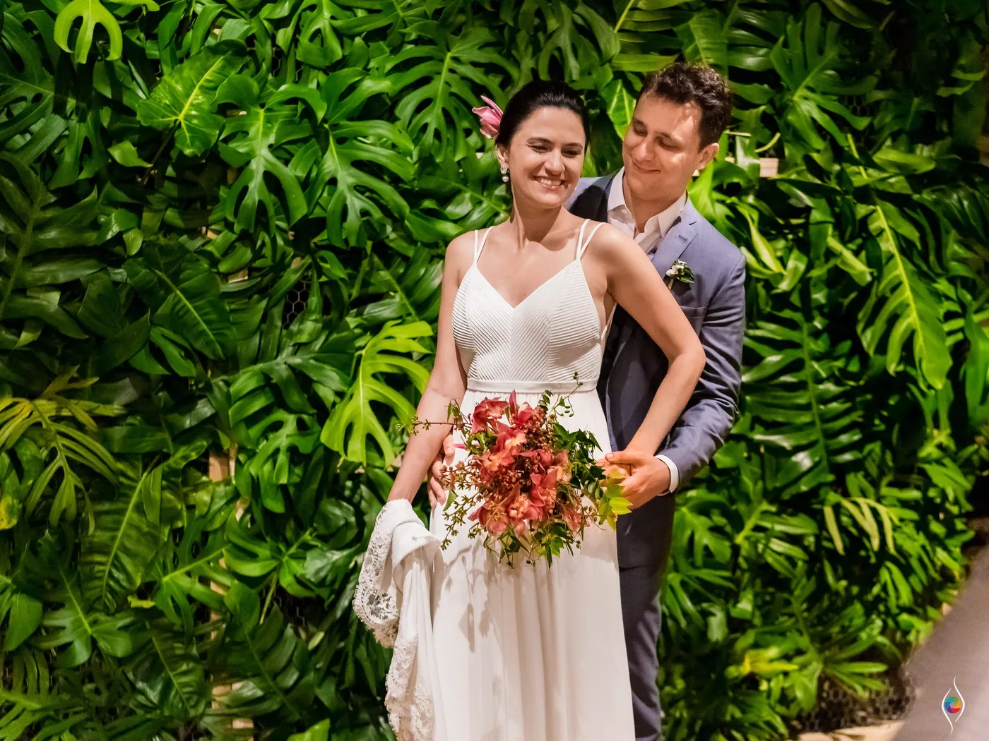 Evy holds a red bouquet as Thiago stands close behind her against a tropical leaf wall