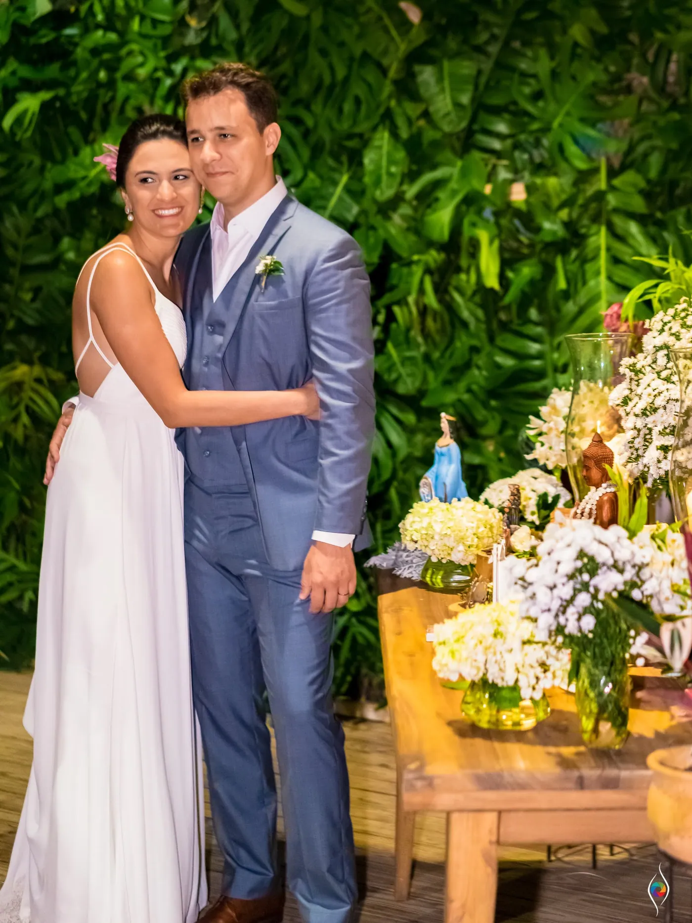 Evy and Thiago embrace beside a table of flowers and a small blue figurine, leaves behind them
