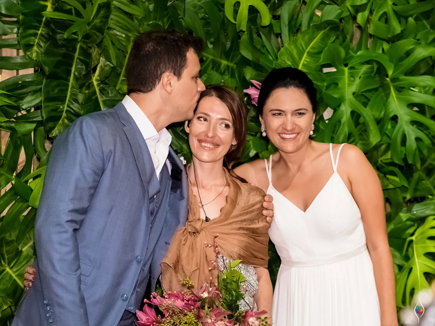 Thiago kisses the cheek of a woman in a brown shawl while Evy laughs beside them at the leaf wall
