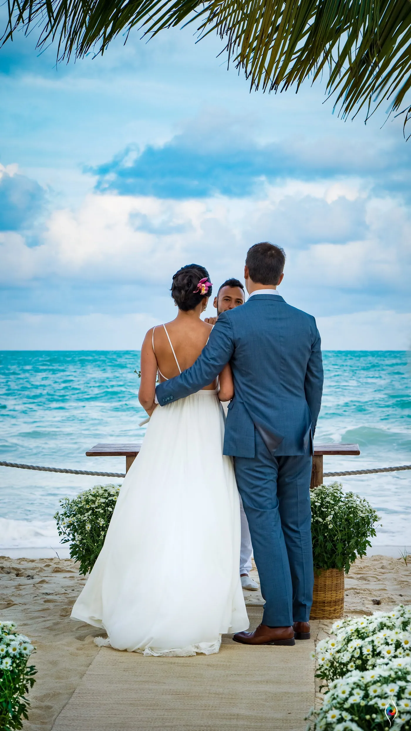 Thiago holds Evy close as they face the sea, white flowers and a palm leaf around them
