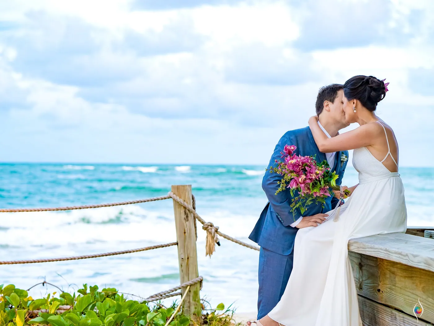 Thiago kisses Evy as they sit by a rope rail, her bouquet in hand and the turquoise sea behind