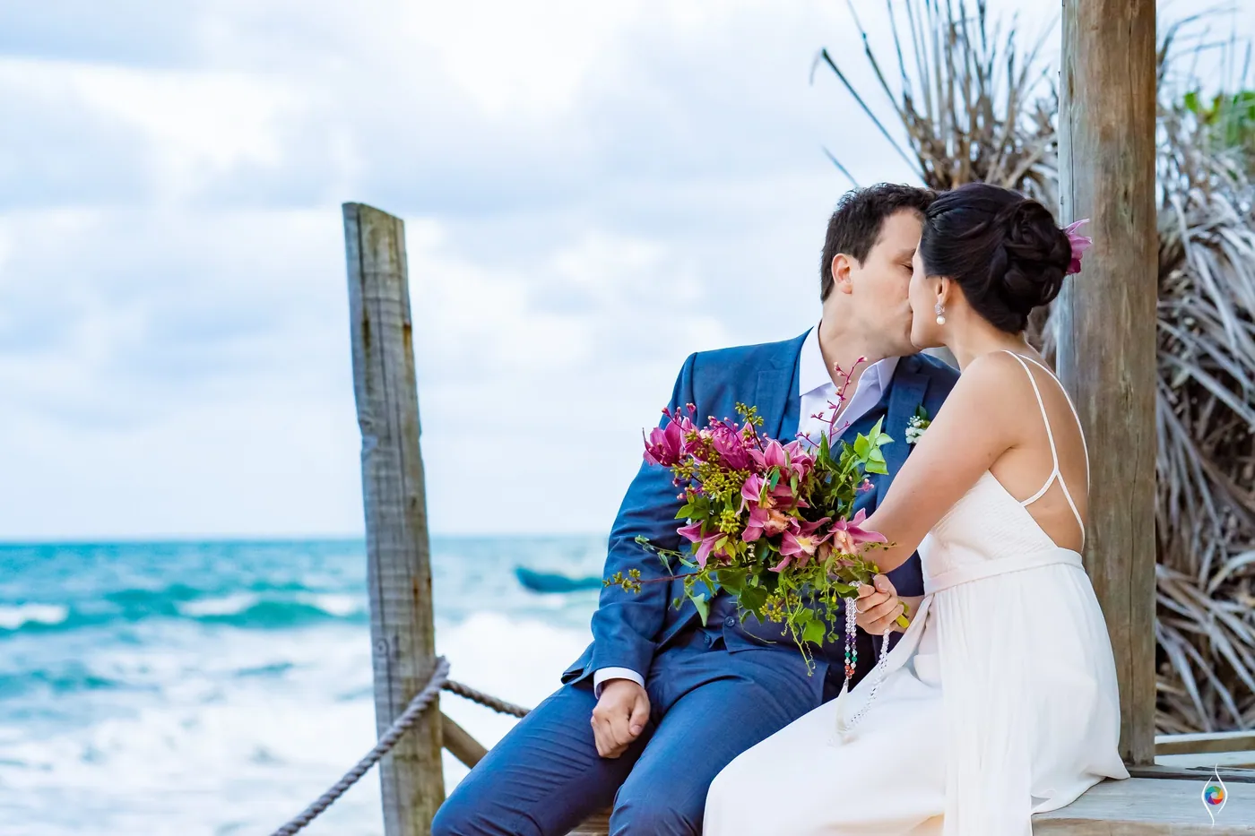Thiago kisses Evy's forehead as they sit close under thatch, the sea glinting beside them