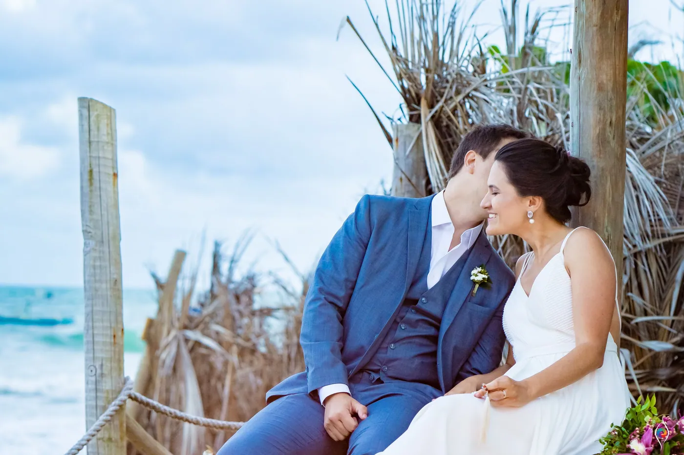 Evy rests her head on Thiago's shoulder and smiles as he kisses her hair, thatch behind them