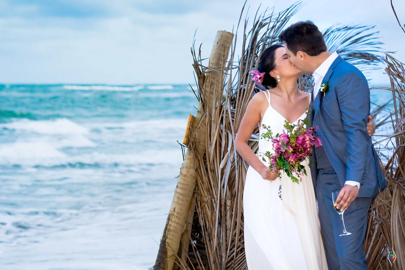 Thiago kisses Evy's cheek as she holds a bouquet and a glass, palm thatch and waves behind them