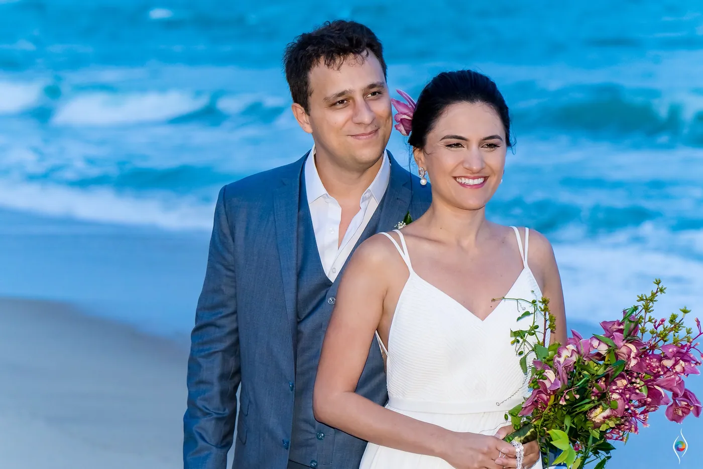 Evy and Thiago smile for the camera on the shore, her pink bouquet in front and surf behind