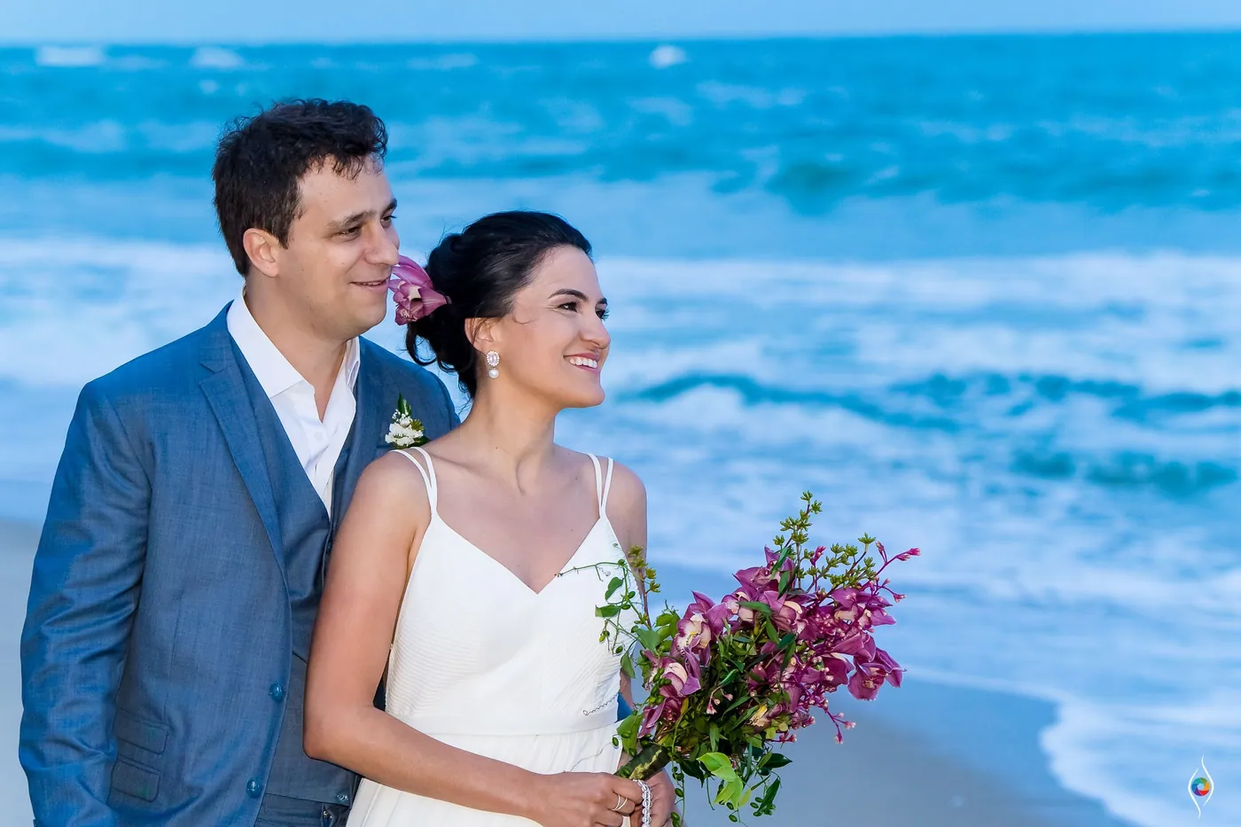 Evy smiles softly past the camera as Thiago stands close, her bouquet held low against the blue surf