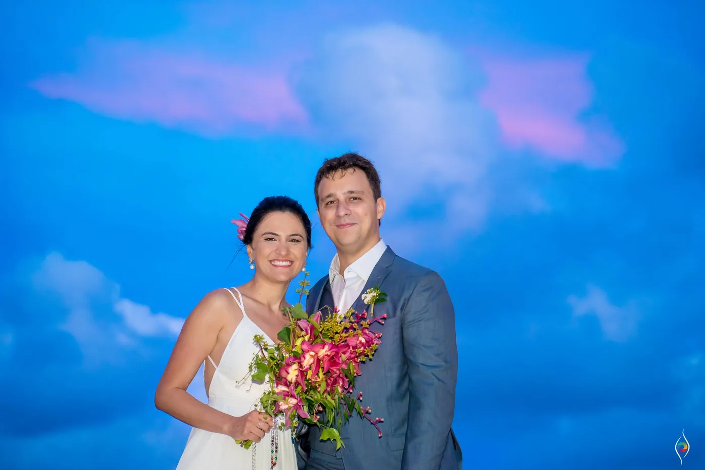 Evy and Thiago pose with her bouquet against a deep blue sky streaked with pink clouds