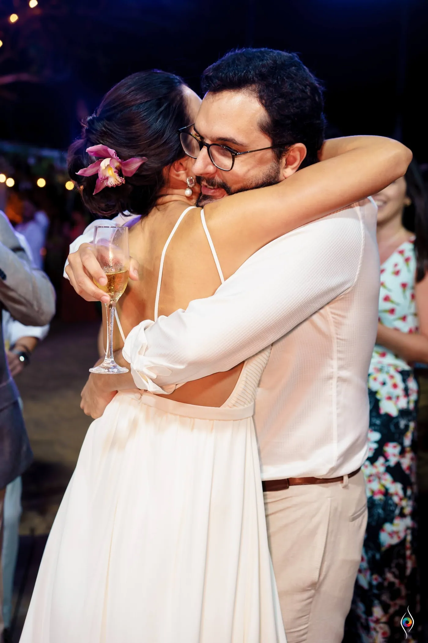 Evy hugs a man in glasses and a white shirt, a drink in her hand, string lights behind them